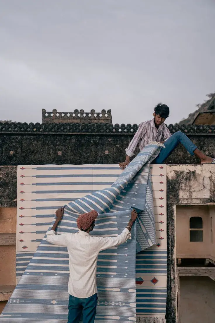 Two men unfolding a large blue and white flatwoven rug in front of an old fort wall